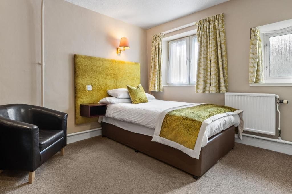 Ein Hotelzimmer mit Bett, Sessel, Nachttisch, Lampe, Fenster mit Vorhängen und Heizkörper.