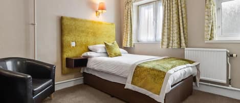 A hotel room with a bed, a chair, a nightstand, a lamp, a window with curtains, and a radiator.