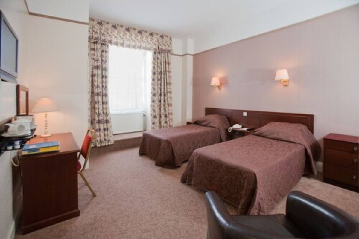 Ein Hotelzimmer mit zwei Betten, einem Schreibtisch mit Lampe, einem Stuhl und einem fenster mit Vorhängen.