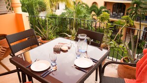 A dining table set with plates, cutlery, and a pitcher on a balcony with wicker chairs and potted plants.