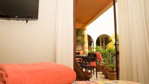 A hotel room with a flat-screen TV mounted on the wall, a bed with an orange blanket, and a view of an outdoor patio with a table, chairs, and potted plants.