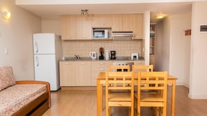 A compact kitchen with wooden cabinets, a microwave, a coffee maker, and a refrigerator. There is a wooden dining table with chairs and a small sofa with a patterned cover. - Civico Apartments (San Carlos de Bariloche)