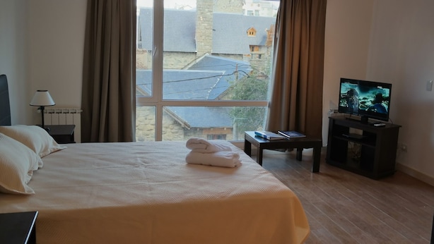 A hotel room with a large bed, a TV on a stand, a bedside table with a book, and a view of buildings outside the window. - Civico Apartments (San Carlos de Bariloche)