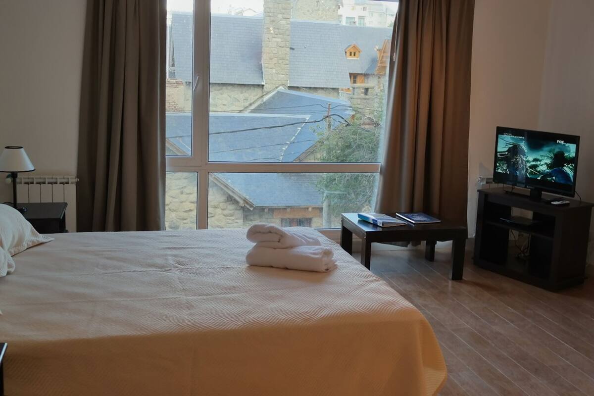 Habitación de hotel con una cama grande, un televisor en un mueble bajo, una mesita de noche con un libro y vistas a edificios al exterior.