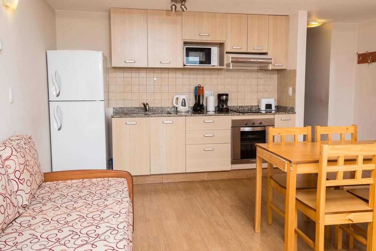 Cocina pequeña con armarios de madera, microondas, cafetera y estufa. Hay una mesa de comedor de madera con sillas.