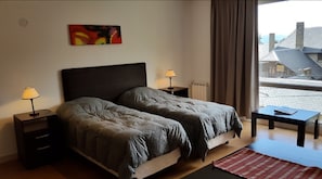Quarto com duas camas, luminárias de cabeceira, radiador, janela com cortinas e vista para edifícios lá fora.