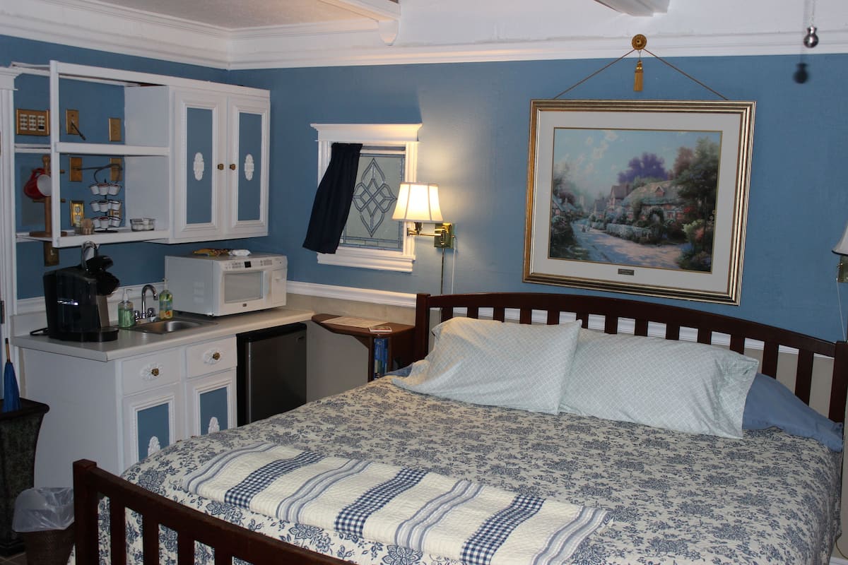 A bedroom with a bed, a nightstand, a lamp, a painting, and a kitchenette.