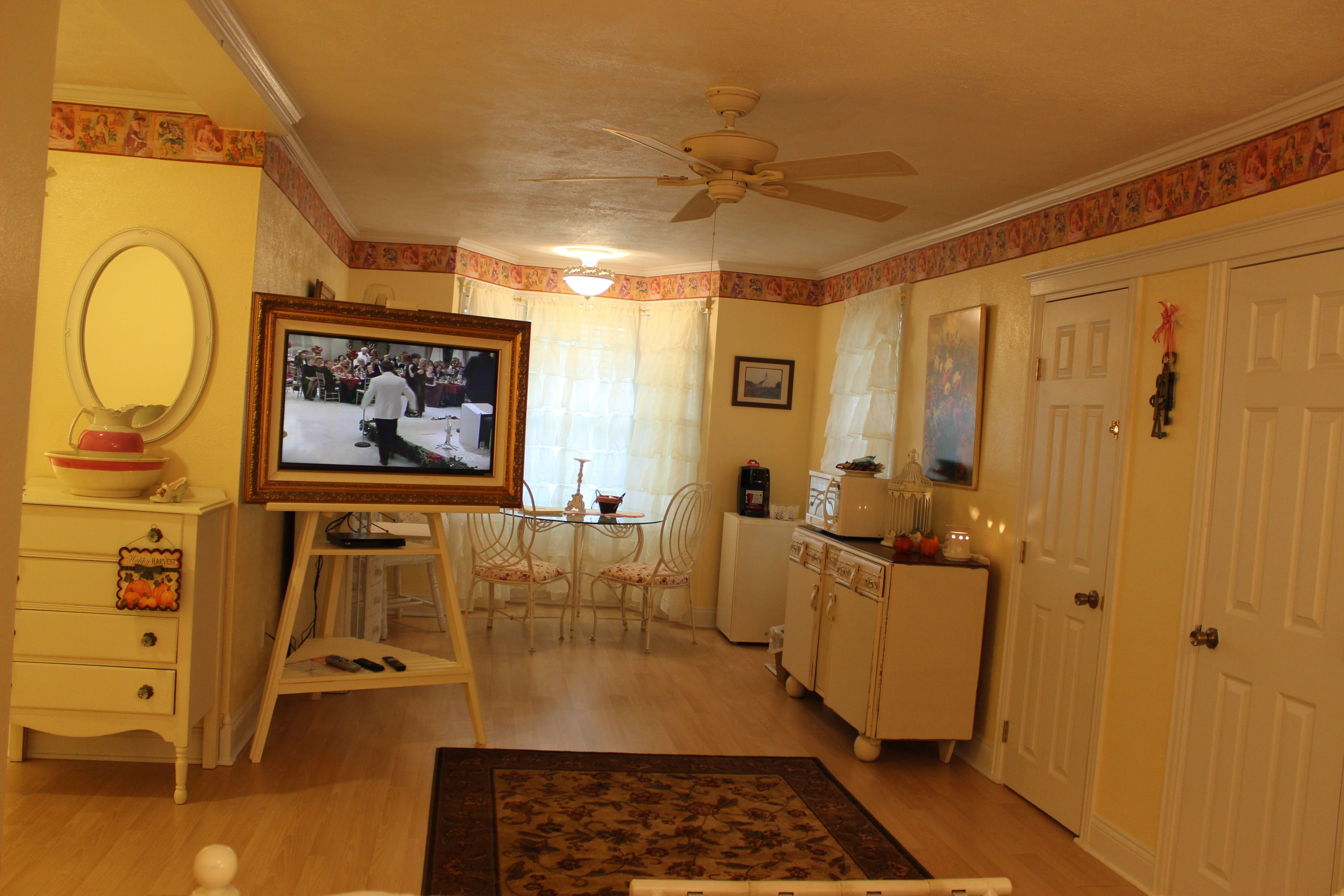 Una sala de estar con televisor sobre un mueble bajo, una mesa de comedor con sillas, un armario blanco y un ventilador de techo.