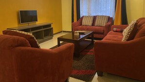 A living room with a sofa set, a coffee table, a flat-screen TV, and a window with curtains. - Milimani Serviced Apartments (Nakuru)