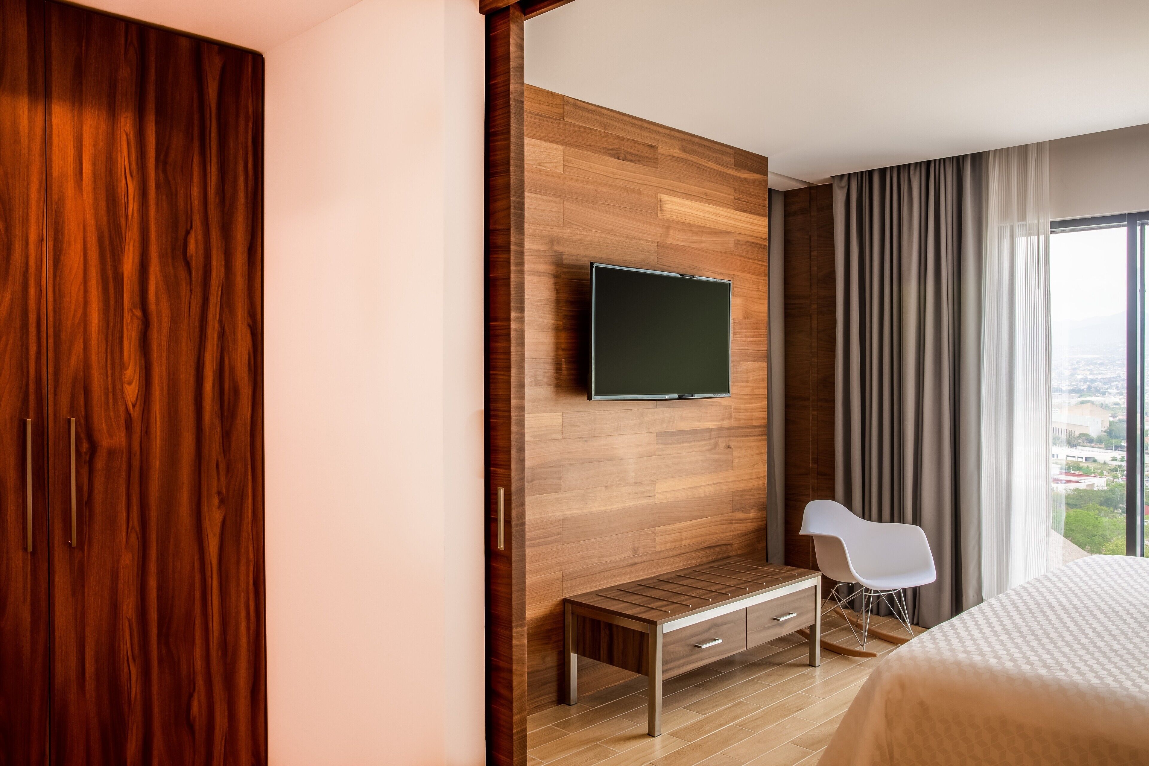 Una habitación de hotel moderna con una pared de madera, un televisor de pantalla plana sobre un banco de madera, una silla blanca y una cama grande.
