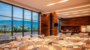 A modern dining area with tables set for meals, large windows offering a view of the city and mountains, and a wooden accent wall. - Fiesta Inn Tuxtla Fashion Mall (Tuxtla Gutierrez)