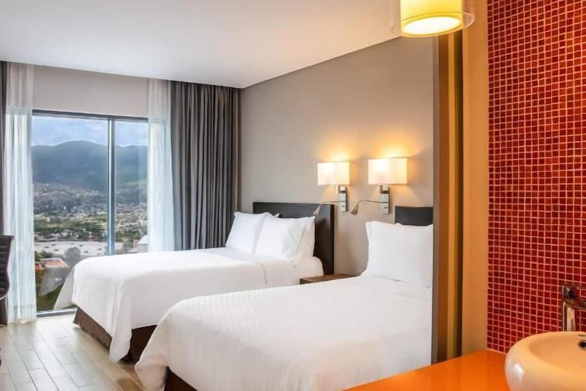 Ein Hotelzimmer mit zwei Betten, einem Schreibtisch, einem Stuhl und Blick auf eine Stadtlandschaft. Das Badezimmer hat rote Fliesen.