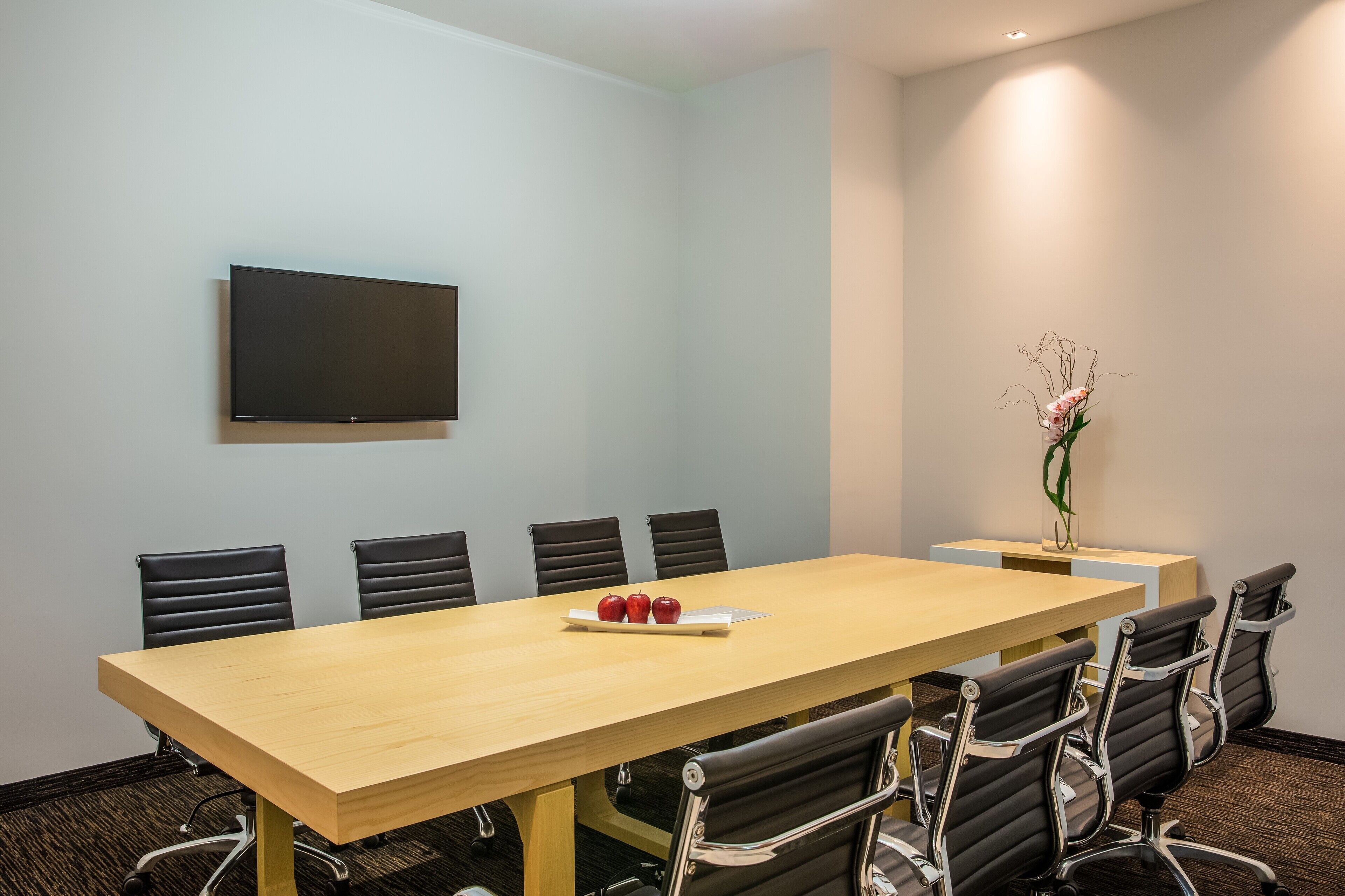 A conference room with a long table, six chairs, a flat-screen TV, and a vase with flowers on a side table.
