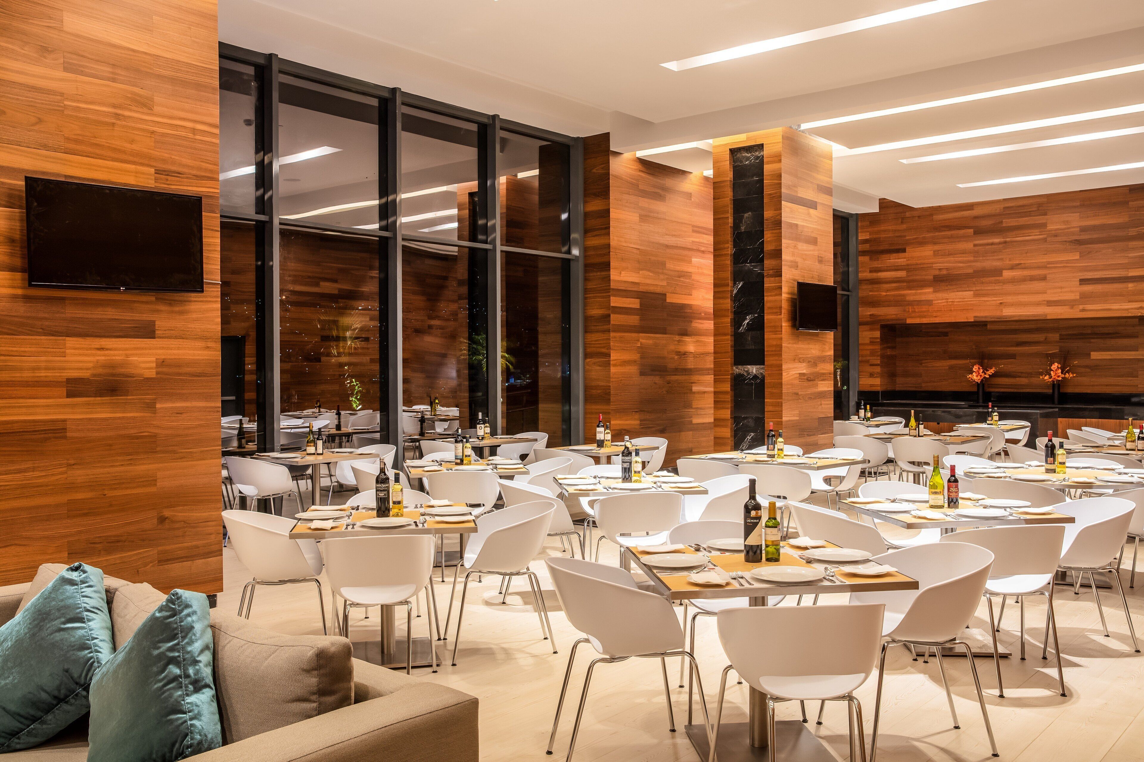 Un área de comedor moderna con sillas y mesas blancas, paneles de madera en las paredes y un televisor de pantalla plana montado en la pared.