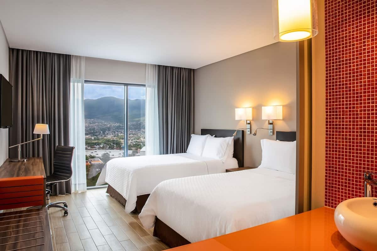 Ein Hotelzimmer mit zwei Betten, einem Schreibtisch, einem Stuhl, einem Fernseher und Blick auf die Stadt.