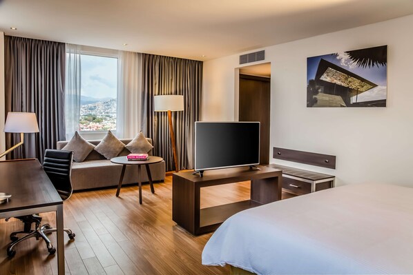 Habitación de hotel con cama, escritorio con televisor, sofá con vistas y ventana con cortinas.
