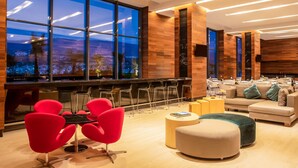 A modern lounge area with a bar, red chairs, a sofa, and large windows offering a city view. - Fiesta Inn Tuxtla Fashion Mall (Tuxtla Gutierrez)