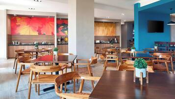 A modern dining area with wooden tables and chairs, a kitchen in the background, and a blue accent wall.