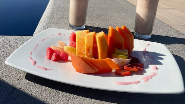 A dining table with a plate of fruit and two drinks, set outdoors with a pool and building in the background. - Fairfield Inn & Suites Aguascalientes (Aguascalientes)