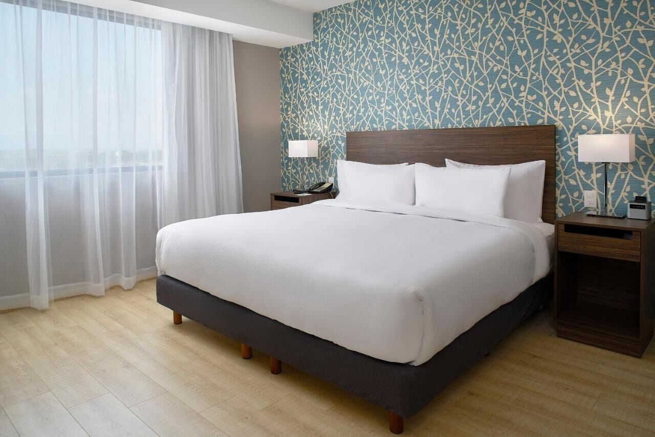 A hotel room with a large bed, two bedside tables, a nightstand, a window with sheer curtains, and a patterned wall.