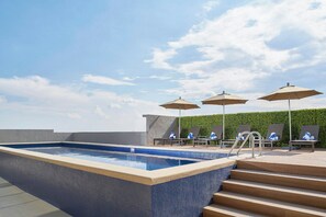Una piscina en la azotea con borde de azulejos azules, rodeada de sillones de descanso y sombrillas, y un seto verde al fondo.