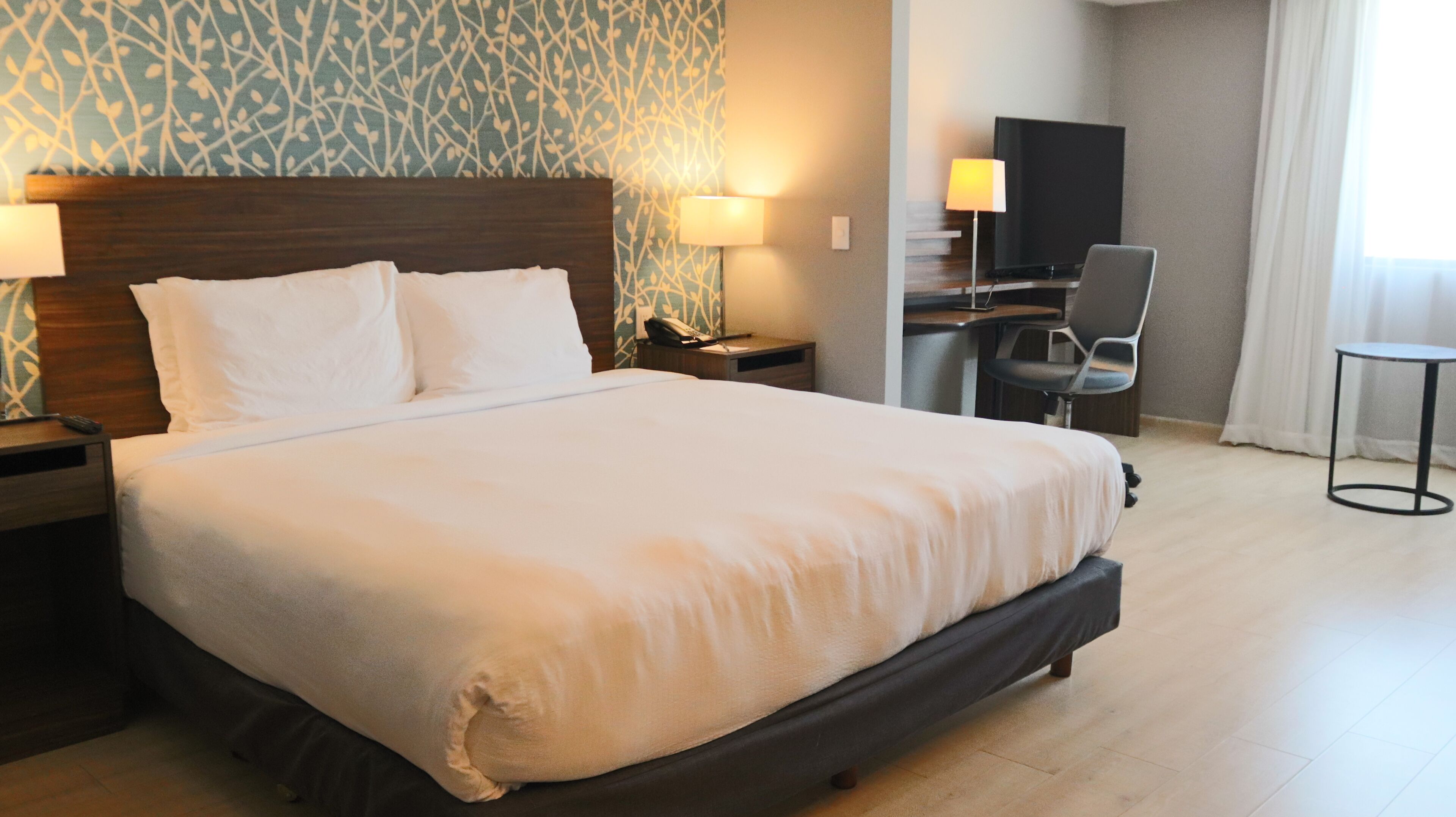 A hotel room with a large bed, a desk with a chair, a TV, and a lamp.