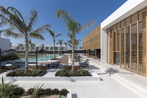 Un área junto a la piscina con sillones de descanso, palmeras y la fachada moderna de un edificio.
