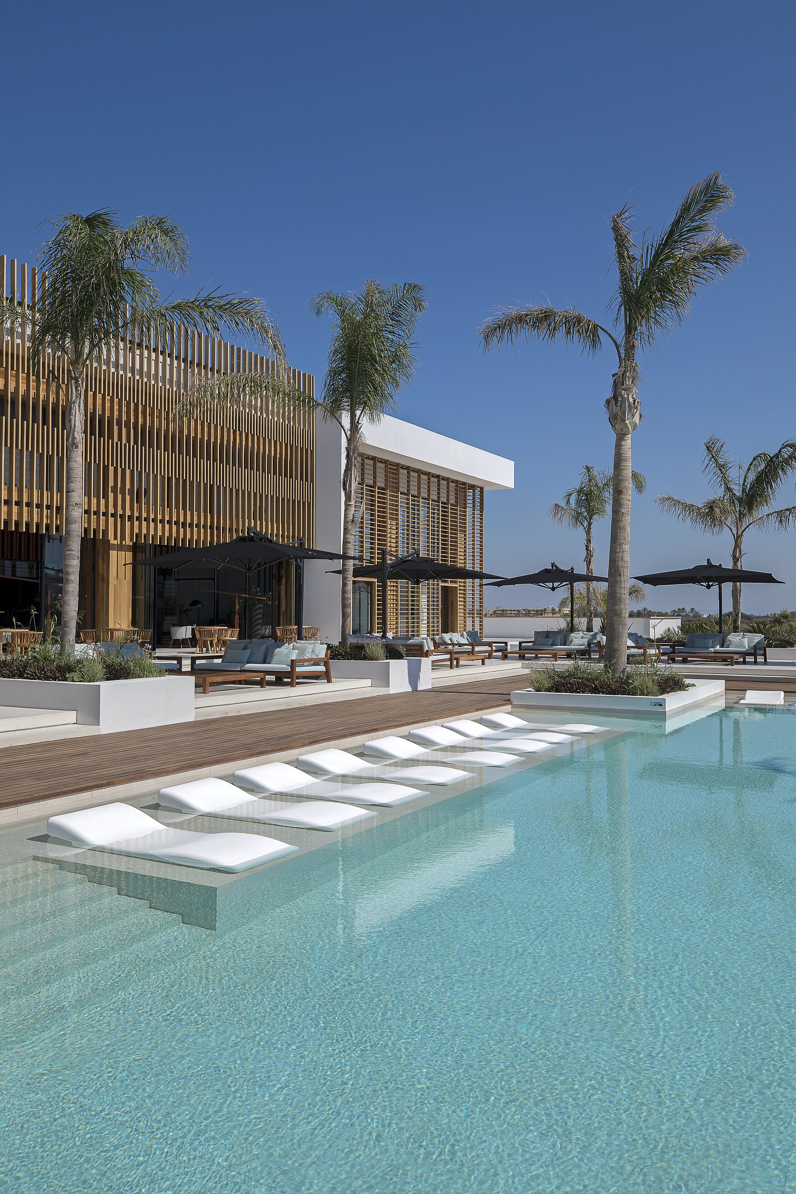 Un área junto a la piscina con sillones de descanso y sombrillas, un edificio moderno con listones verticales de madera y palmeras.