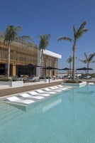 Un área junto a la piscina con sillones de descanso y sombrillas, un edificio moderno con listones verticales de madera y palmeras.