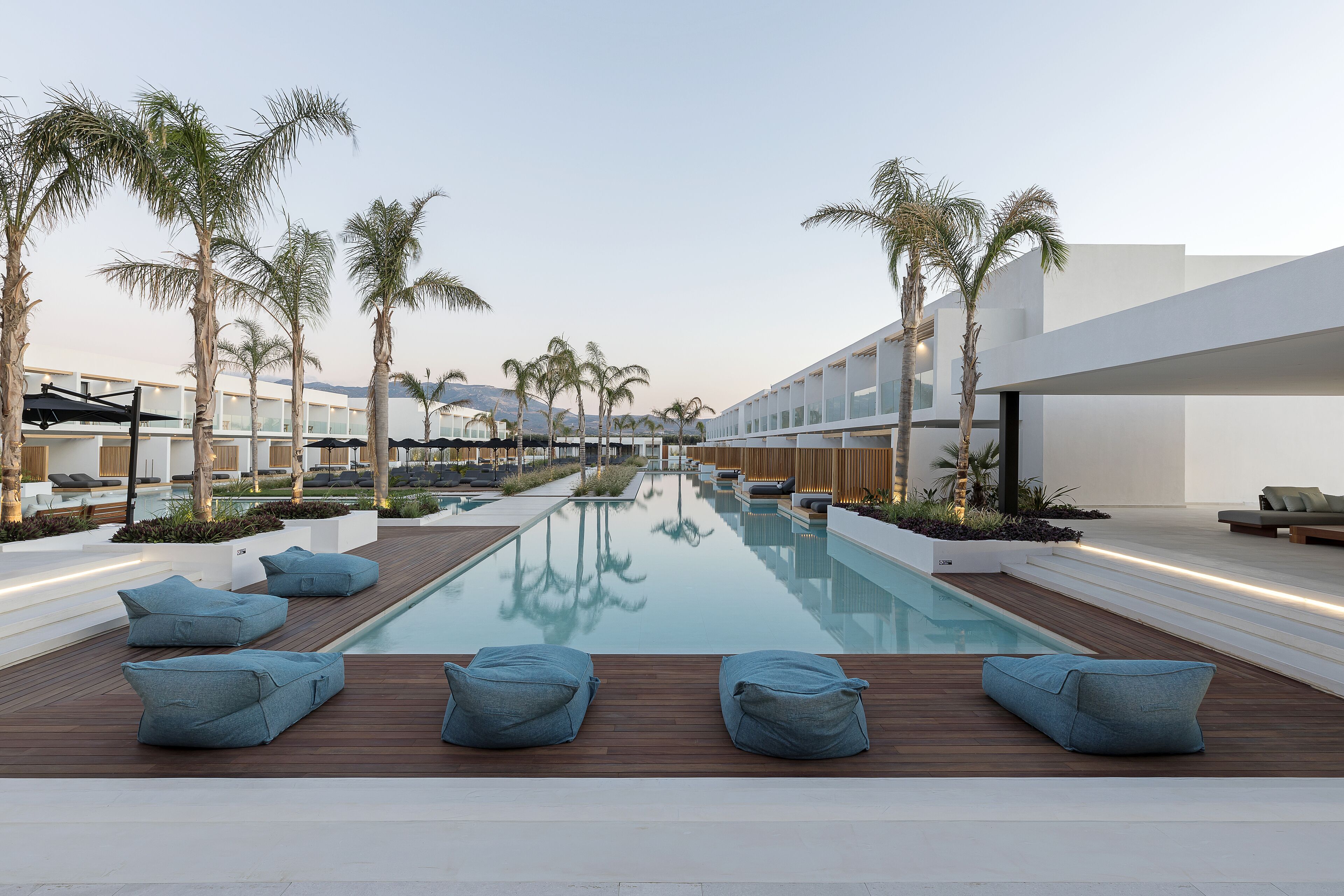 A modern pool area with wooden decking, blue bean bags, and palm trees.
