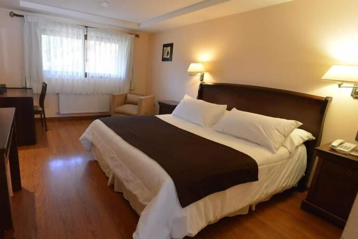 Habitación de hotel con cama, mesitas de noche, escritorio, silla y ventana con cortinas.