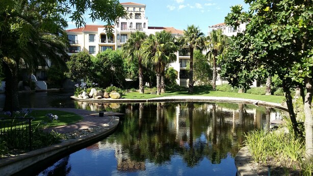 A serene courtyard with a reflective pond, surrounded by lush greenery and multi-story buildings with balconies. - Luxurious Apartment in Century City (Cape Town)
