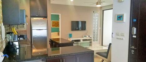 A modern kitchen with a refrigerator, stove, and dining area. There is a TV mounted on the wall and a window providing natural light.