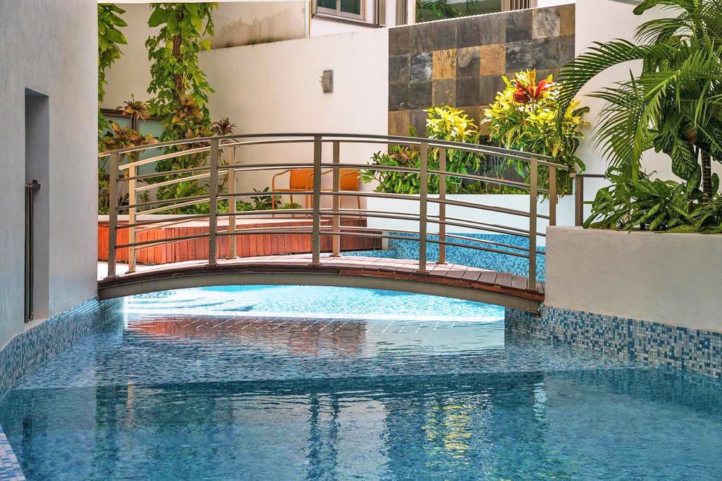 An indoor swimming pool with a wooden bridge, surrounded by plants and a tiled wall.