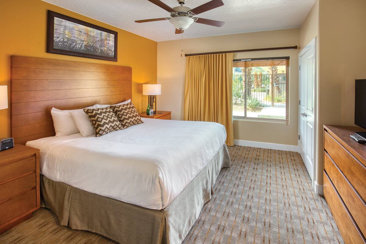 Quarto de hotel com cama grande, cabeceira de madeira, duas mesas de cabeceira com luminárias, ventoinha no teto, televisão em cômoda de madeira e janela com cortinas.