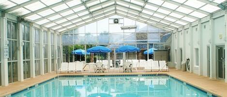 An indoor swimming pool with clear glass walls, blue umbrellas, and lounge chairs.