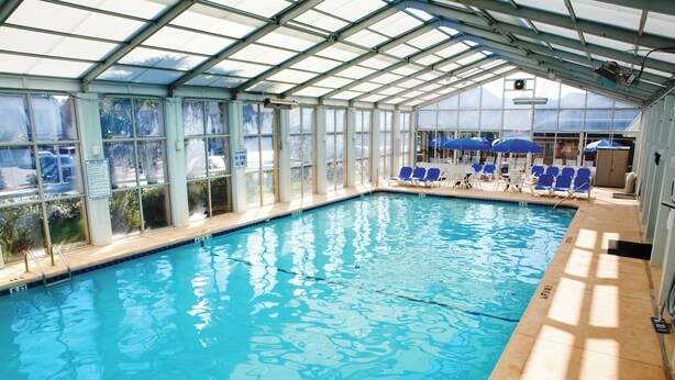 An indoor swimming pool with clear glass walls, blue chairs, and umbrellas. - Club Wyndham Grand Palms (Surfside Beach)