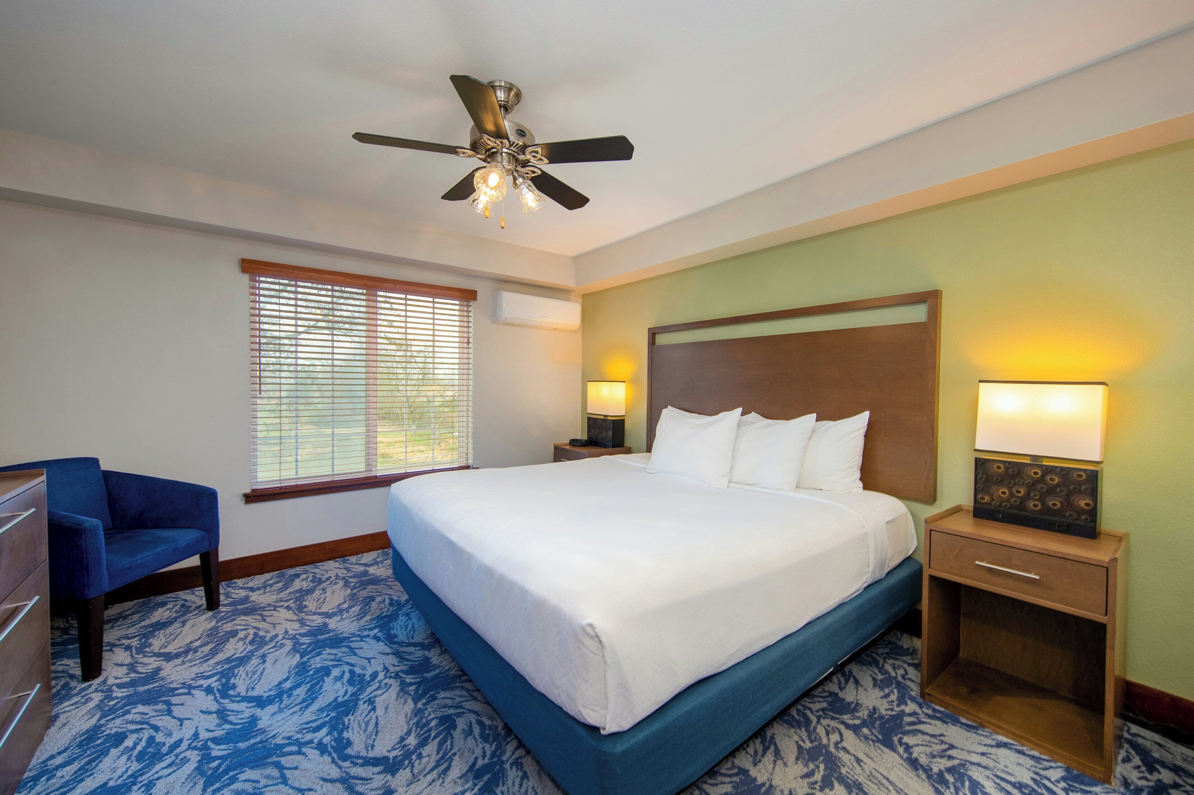 A hotel room with a large bed, a ceiling fan, a nightstand with a lamp, a blue chair, and a window with blinds.
