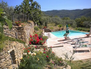 A pool area with a stone wall, a variety of flowers, and a view of hills in the background. - Agriturismo Casale della Torre (Cortona)