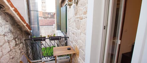 A narrow balcony with a wooden table and chairs, a potted plant, and a view of a tower in the background.
