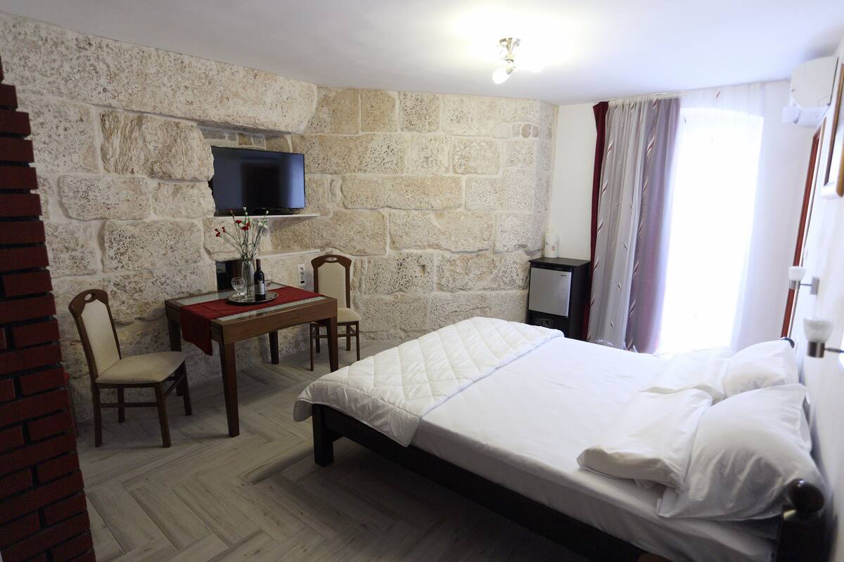 A hotel room with a stone wall, a flat-screen TV, a wooden desk with chairs, a bed with white linens, and a window with curtains.