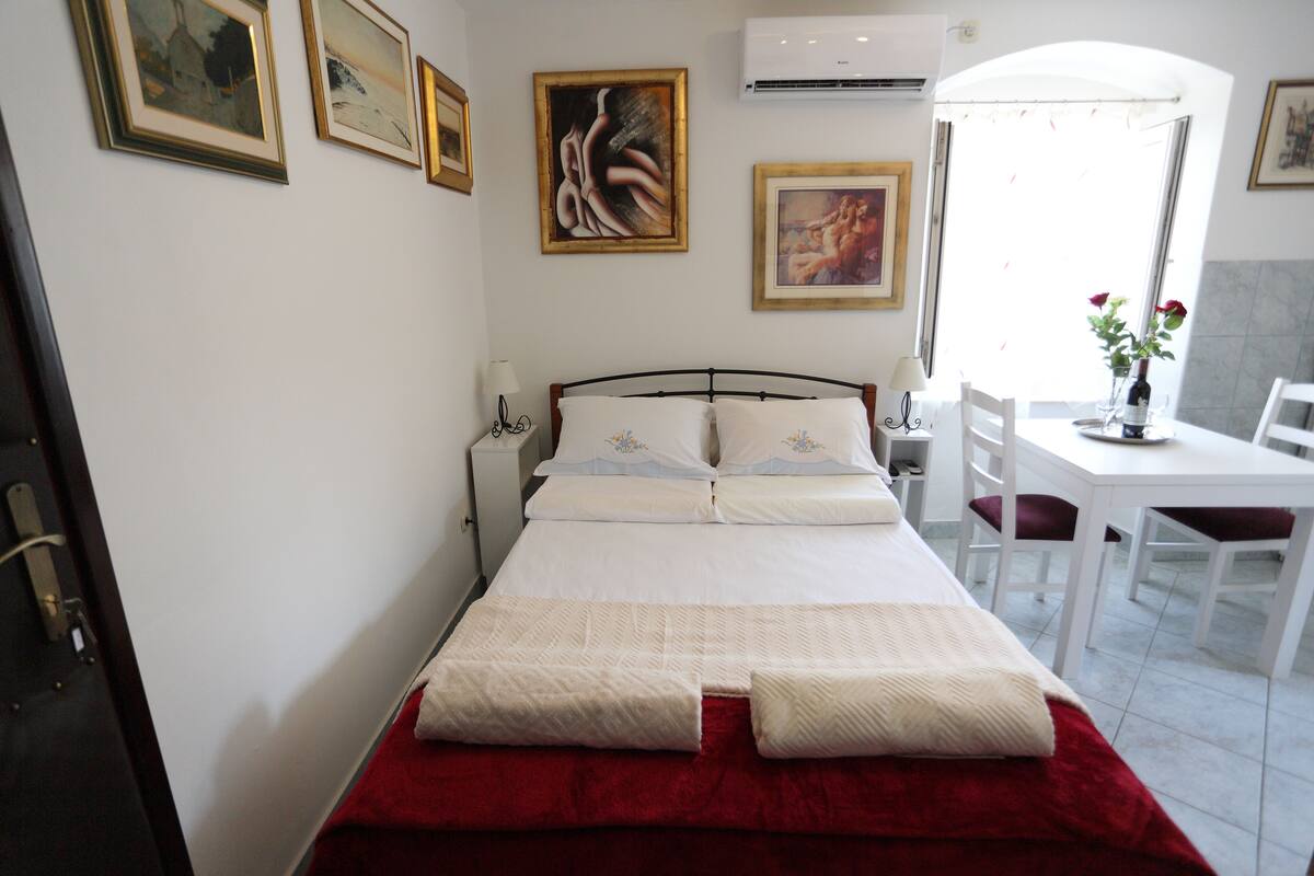 A neatly made bed with white linens and a red blanket, a bedside table with a lamp, and a dining area with a white table and chairs.