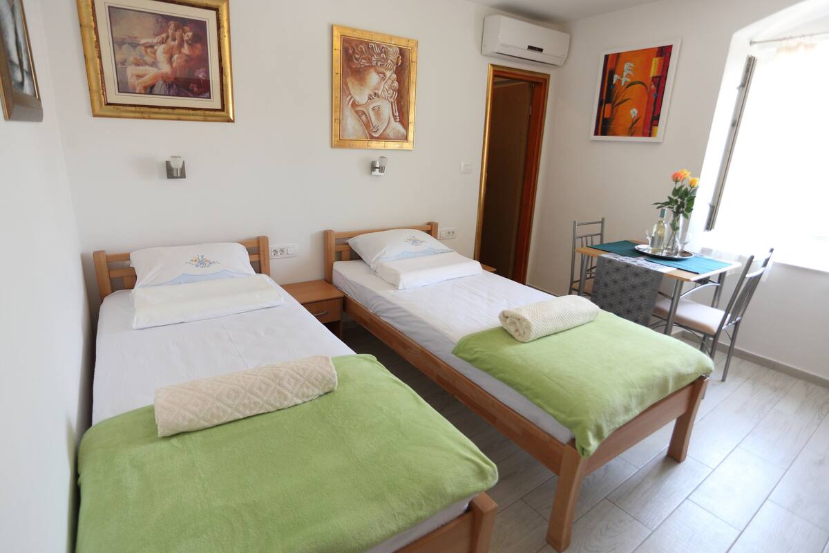 A room with two wooden beds, each with a green blanket and a white pillow. There are paintings on the walls and a window with curtains.