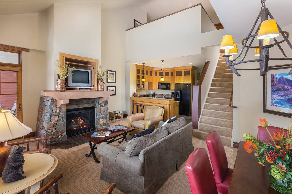 Amplia sala de estar con chimenea, una cocina al fondo y una escalera que sube a un nivel superior.