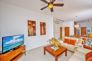 A modern living room with a flat-screen TV, a ceiling fan, a dining area with wooden furniture, and abstract wall art.