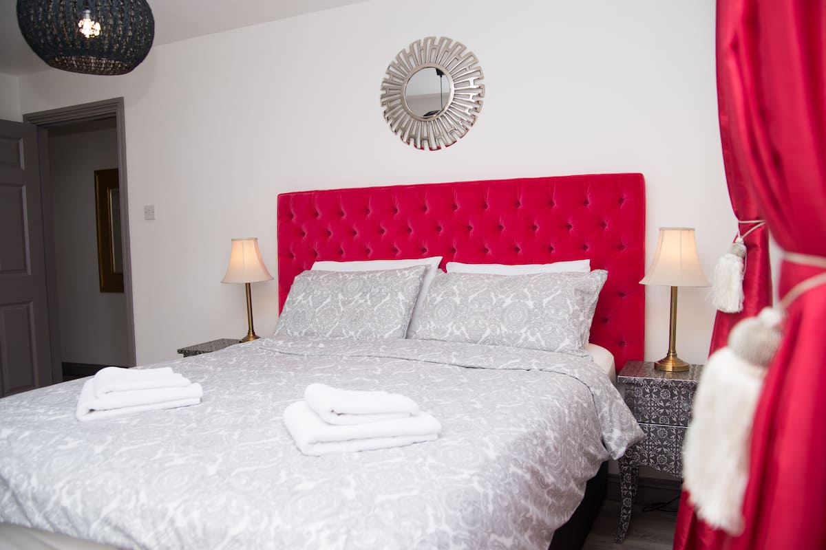 A neatly made bed with white linens and pillows, a red upholstered headboard, two bedside lamps, and a decorative mirror on the wall.