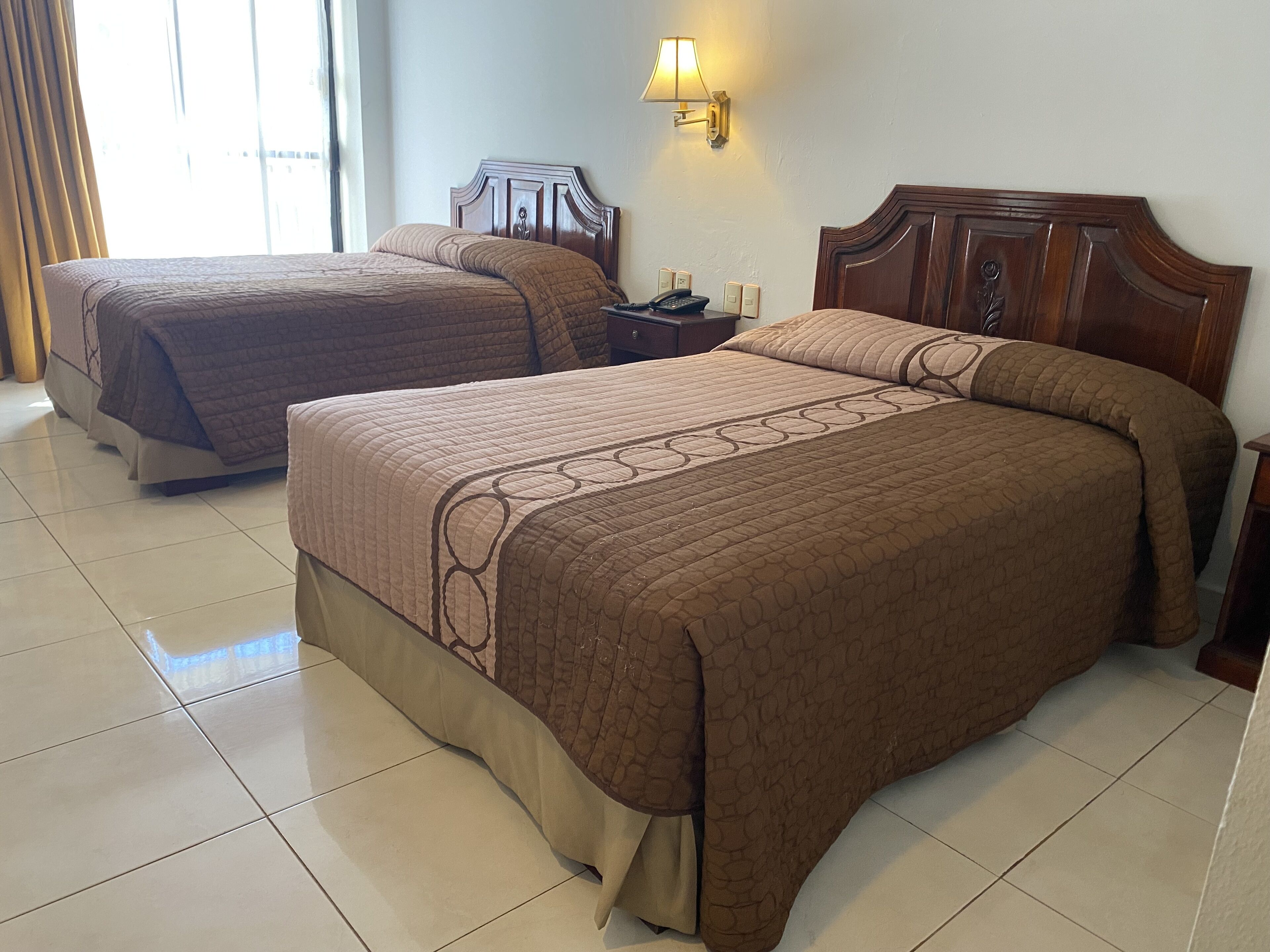 Two double beds with brown bedding, a nightstand with a lamp, and a wall-mounted light fixture.