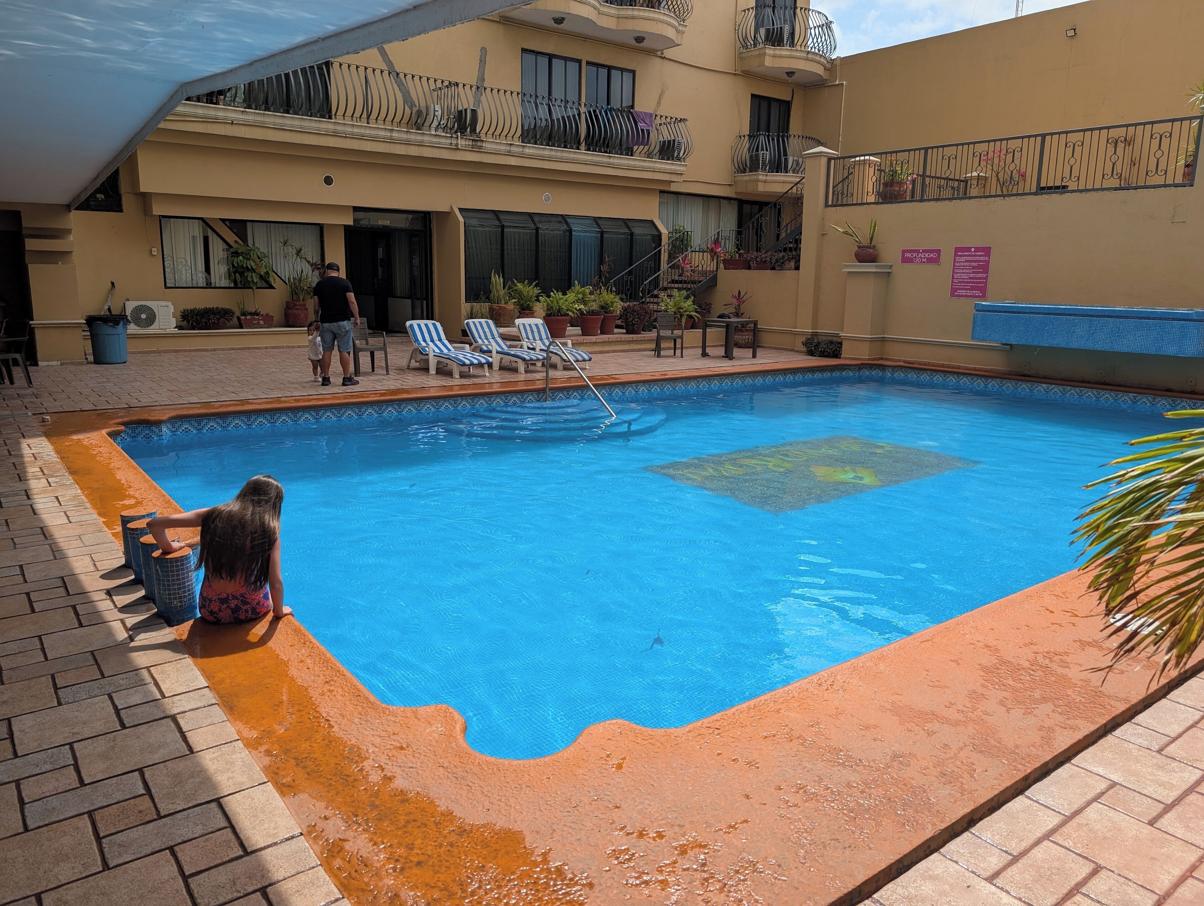 A swimming pool with blue water, surrounded by a brick border. There are lounge chairs and potted plants around the pool.