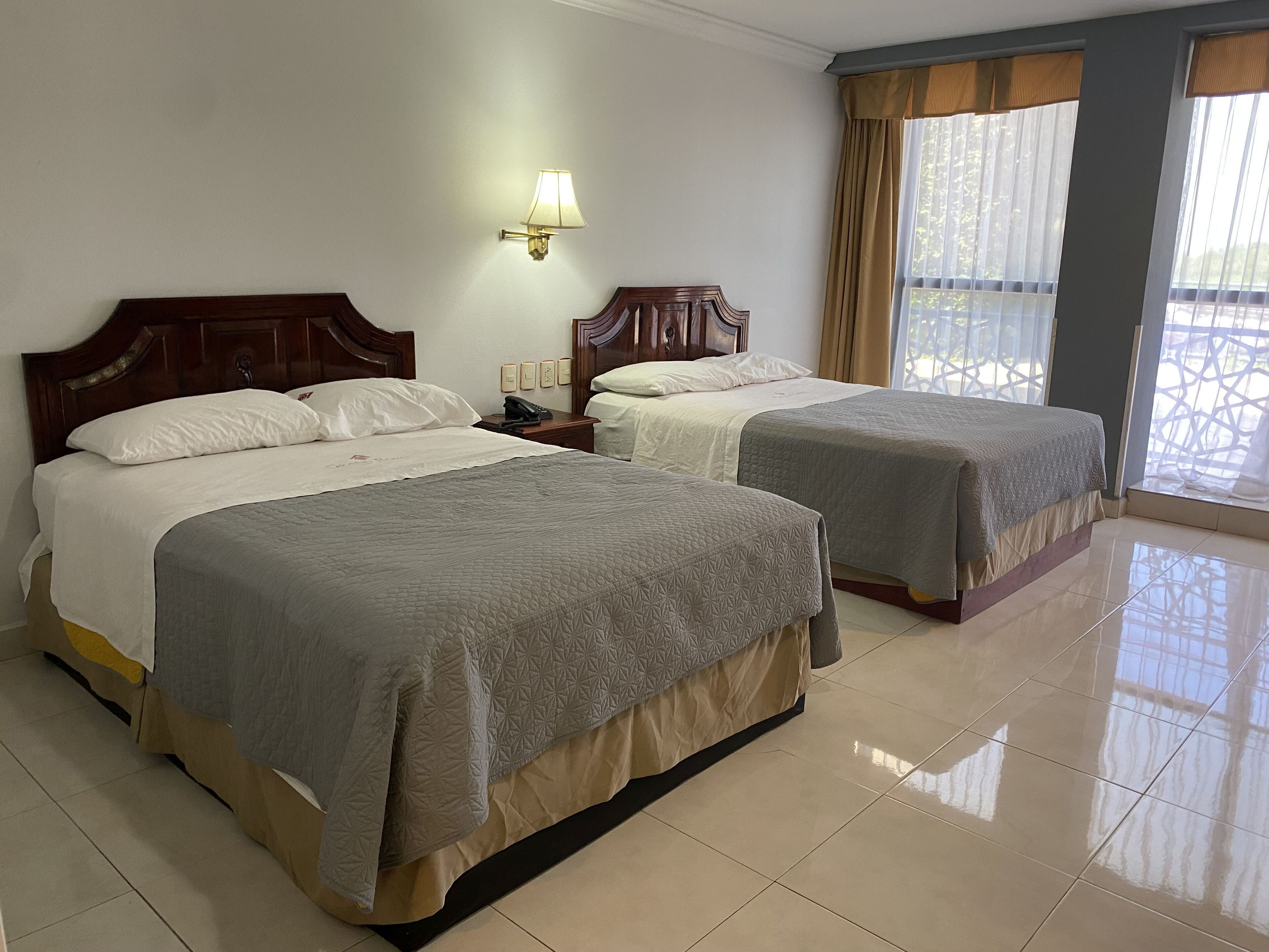 A hotel room with two beds, a nightstand, a lamp, and a window with curtains.