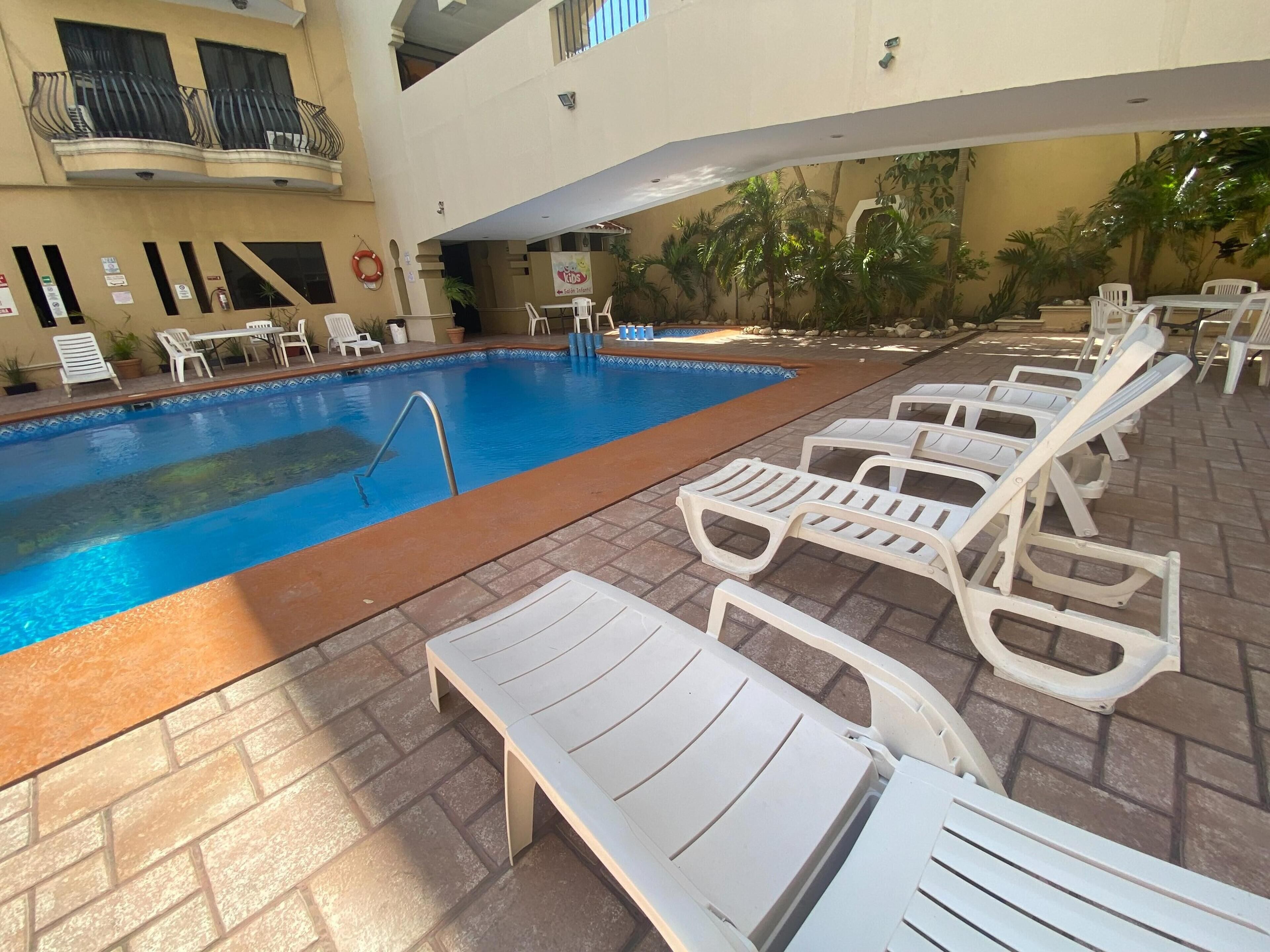 A swimming pool with lounge chairs and a small table, surrounded by potted plants and a building with balconies.